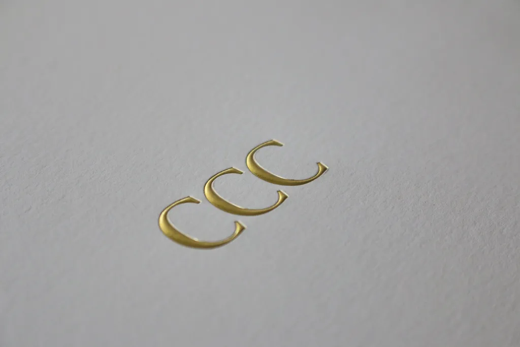 Three gold foil letter Cs stamped and embossed into white cotton paper
