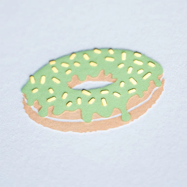 Letterpress donut printed in green and tan ink on white paper, with gold foil sprinkles