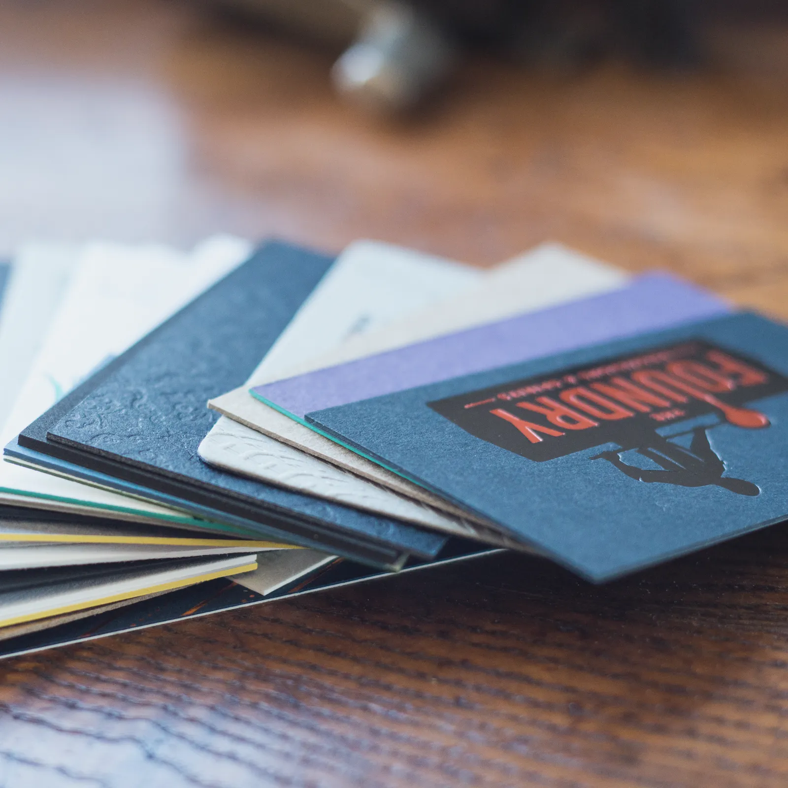 A fan of letterpress business cards on a wooden table — white, cream, black, navy, and lavender stocks with deep debossed patterns, painted edges, and a navy card printed in red and black ink