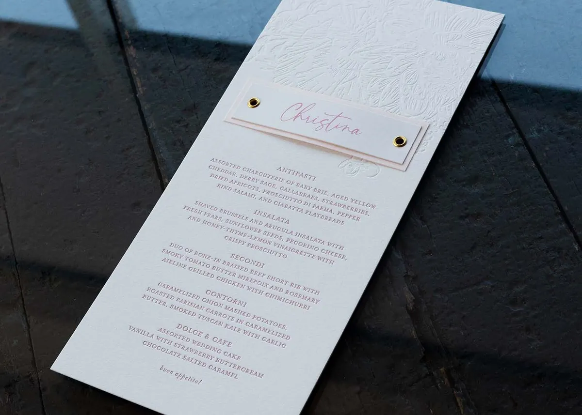 Top view of letterpress wedding menu with die-cut name tag attached by brass eyelet on blush paper